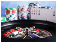 En primer plano se ve el cilindo negro de una ruleta y las casillas rojas y negras. De fondo, varios montones de fichas de casino y los cuatro ases de la baraja francesa colocados en vertical.